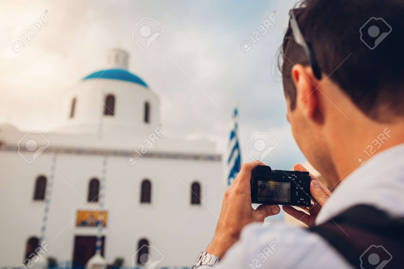 Santorini traveler man taking photo of church with blue dome and flag in Oia, Greece on camera. Tourism, traveling, summer vacation. Traditional architecture - 168610453 Santorini traveler man taking photo of church with blue dome and flag in Oia, Greece on camera. Tourism, traveling, summer vacation. Traditional architecture - 168610453