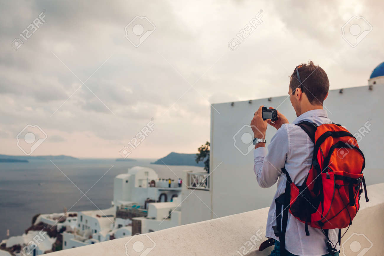 Santorini traveler man taking photo of Caldera from Oia, Greece on camera. Backpacker travels during summer vacation. Tourist admiring Aegean sea landscape. - 168610455 Santorini traveler man taking photo of Caldera from Oia, Greece on camera. Backpacker travels during summer vacation. Tourist admiring Aegean sea landscape. - 168610455