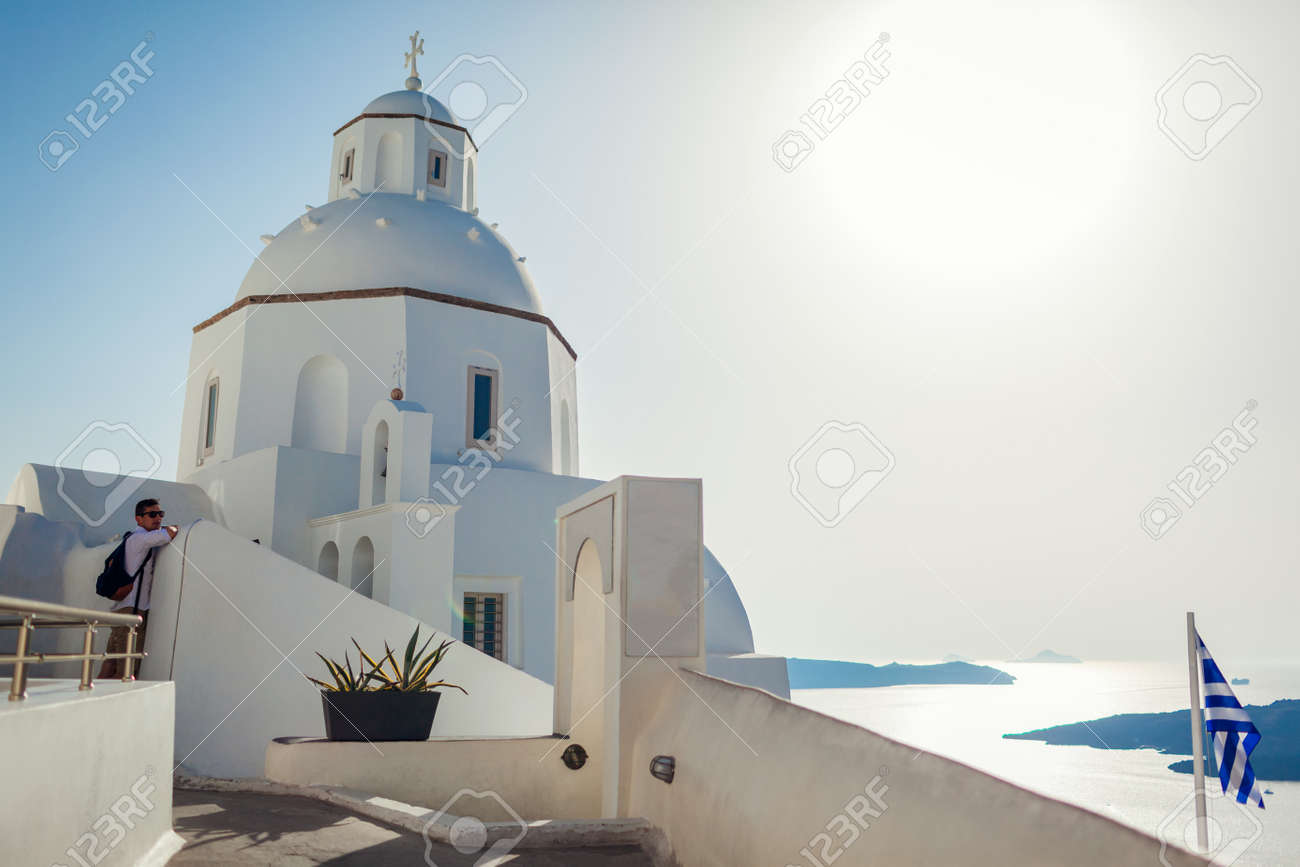 Man tourist enjoying traditional architecture walking by white church on Santorini island with sea landscape in Fira. Greek flag - 171513139 Man tourist enjoying traditional architecture walking by white church on Santorini island with sea landscape in Fira. Greek flag - 171513139