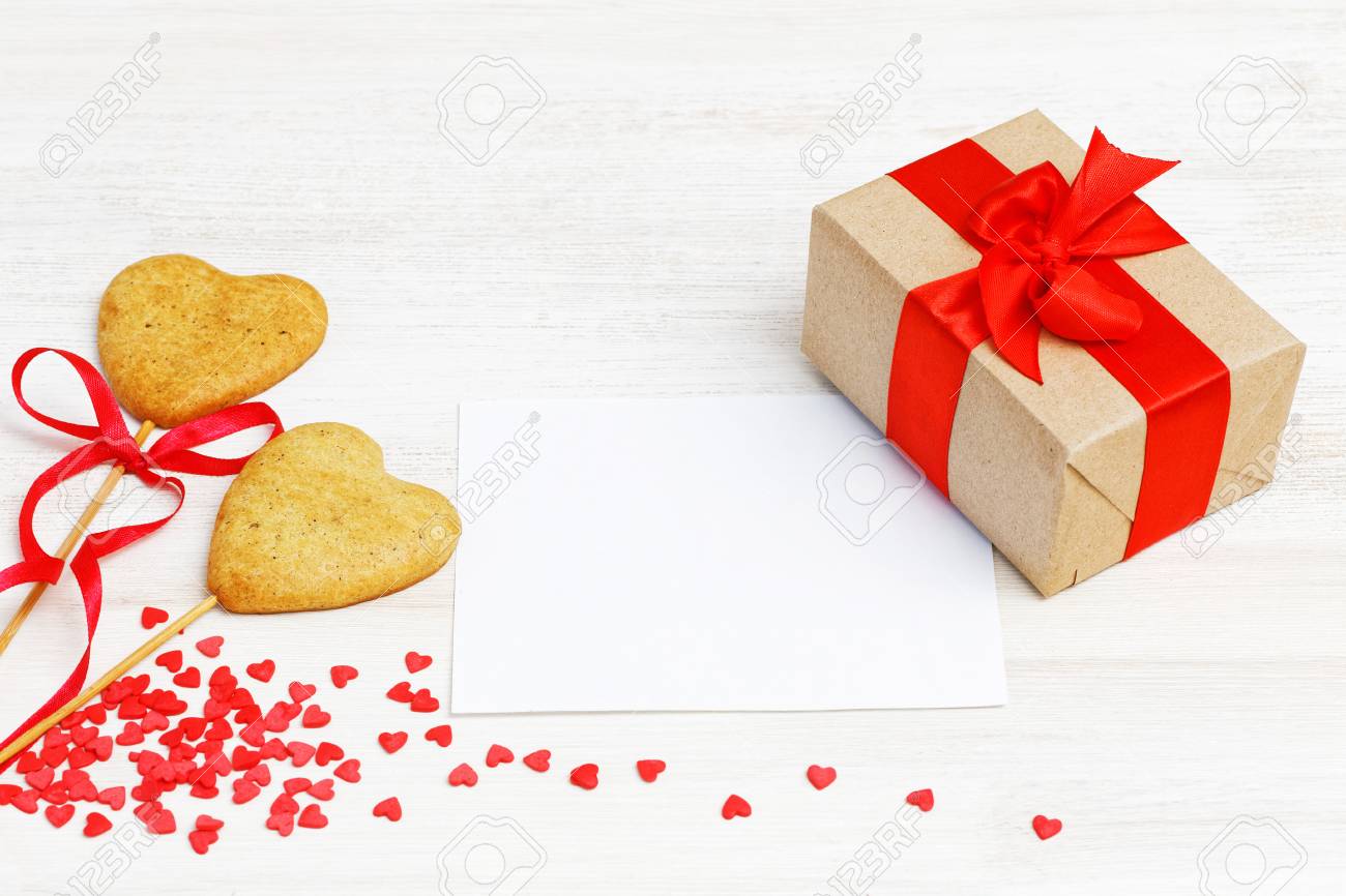 バレンタインデーのテンプレート クラフト紙に包まれたギフト付きのボックス 赤いリボン 白いカード ハート の形をしたスティックに2つの自家製クッキー 赤いハート型の装飾 白い木製の背景 の写真素材 画像素材 Image