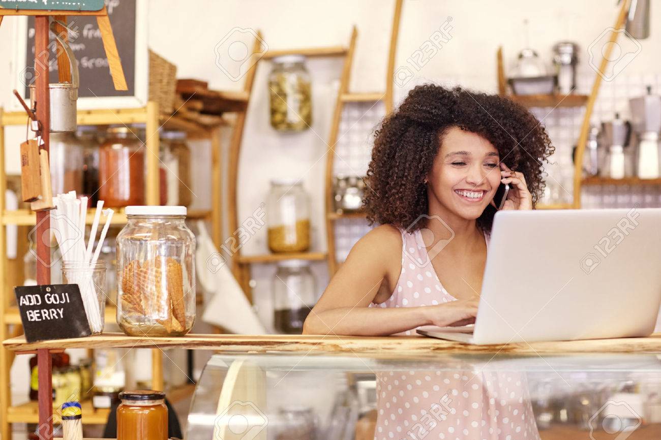 Happy young coffee shop owner talking on he mobile phone while typing on her laptop on the counter - 51356657 Happy young coffee shop owner talking on he mobile phone while typing on her laptop on the counter - 51356657