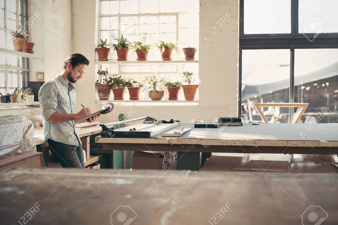 Young male business owner standing in his studio workshop checking figures and stock on his clipboard on a sunny afternoon - 51813672 Young male business owner standing in his studio workshop checking figures and stock on his clipboard on a sunny afternoon - 51813672