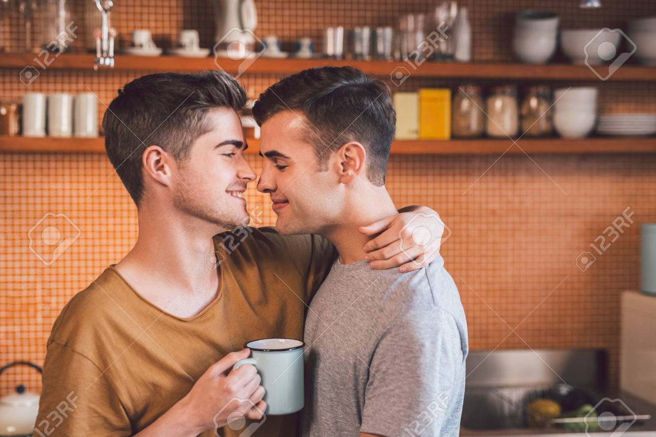 Affectionate Young Gay Couple Standing Arm In Arm Together While Drinking Coffee In Their Kitchen In Affectionate Young Gay Couple Standing Arm In Arm Together While Drinking Coffee In Their Kitchen In