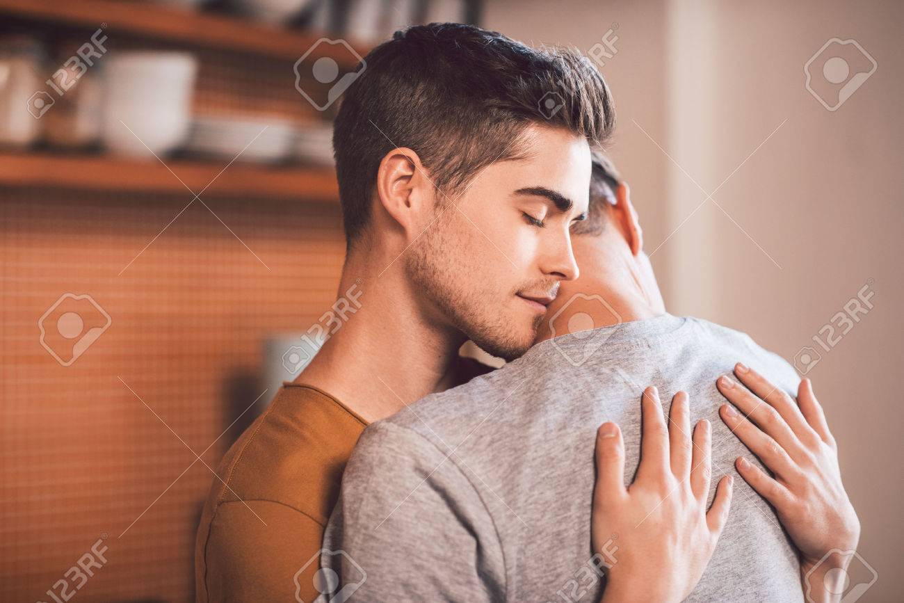 Affectionate Young Gay Couple Hugging Each Other With Their Eyes Closed While Standing In Their Kitchen Affectionate Young Gay Couple Hugging Each Other With Their Eyes Closed While Standing In Their Kitchen