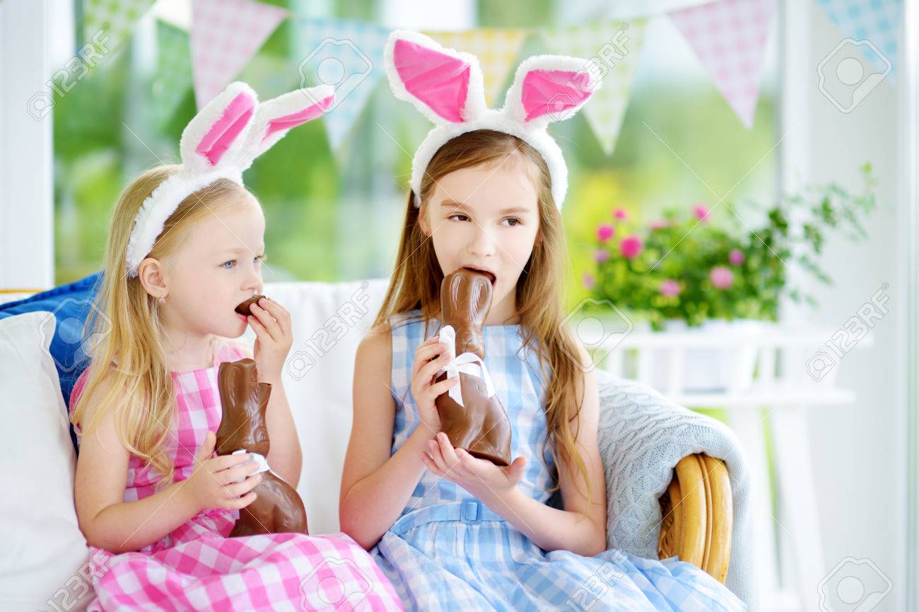 Dos Pequeñas Hermanas Lindas Con Orejas De Conejo Que Comen Los Conejos De  Pascua De Chocolate. Niños Jugando En La Búsqueda De Huevos De Pascua.  Niños Adorables Que Celebran La Pascua En