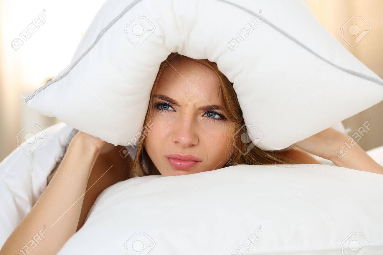 Young beautiful woman lying in bed suffering with insomnia covering head and ears with pillow and making unpleasant face. Noisy neighbour, stress, alarm sound, prevent from sleep concept - 46189010 Young beautiful woman lying in bed suffering with insomnia covering head and ears with pillow and making unpleasant face. Noisy neighbour, stress, alarm sound, prevent from sleep concept - 46189010