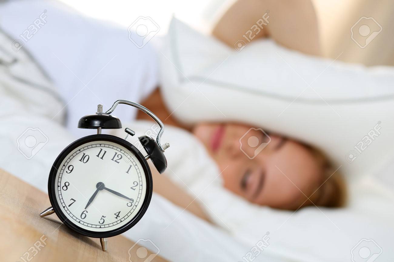 Réveil debout sur une table de chevet va sonner alors que la tête de couverture femme et les oreilles avec l'oreiller faisant visage désagréable. Réveil debout sur une table de chevet va sonner alors que la tête de couverture femme et les oreilles avec l'oreiller faisant visage désagréable.