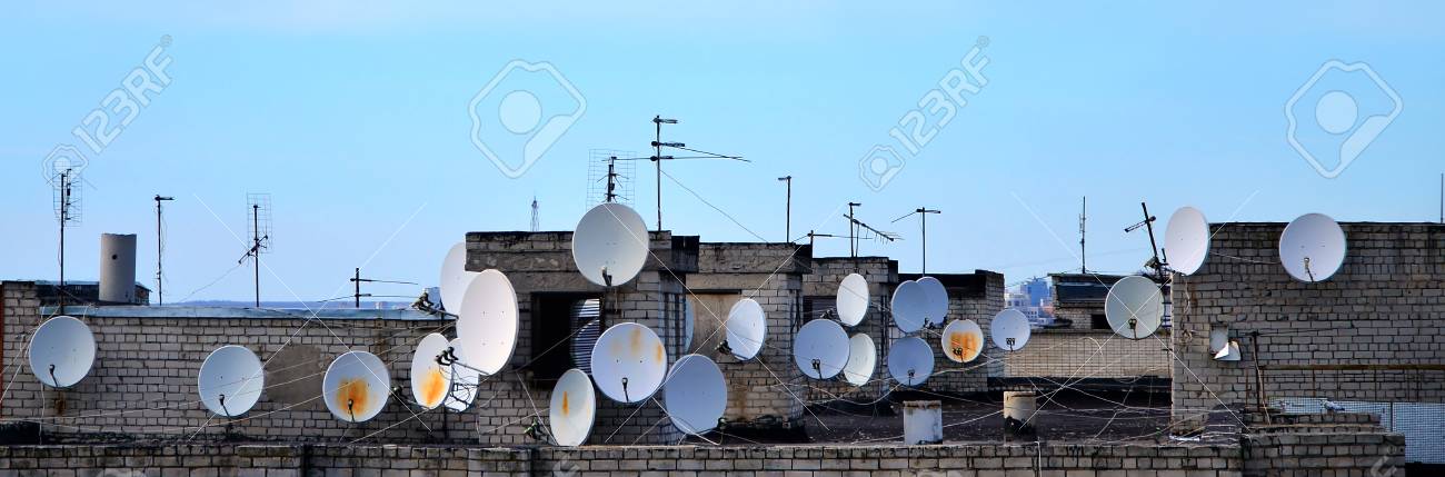 Muchas Antenas De Televisión Satelital Y Por Cable Se Encuentran En La Azotea De Un Edificio De Varias Plantas Bajo Un Cielo Azul. Antiguas Antenas De Televisión En Blanco A La
