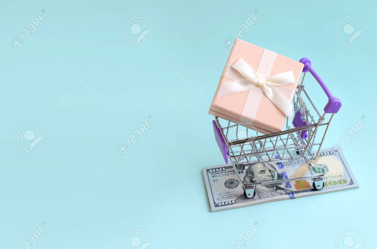 Gift Box In A Small Shopping Cart Lies On A Dollar Bills On Light