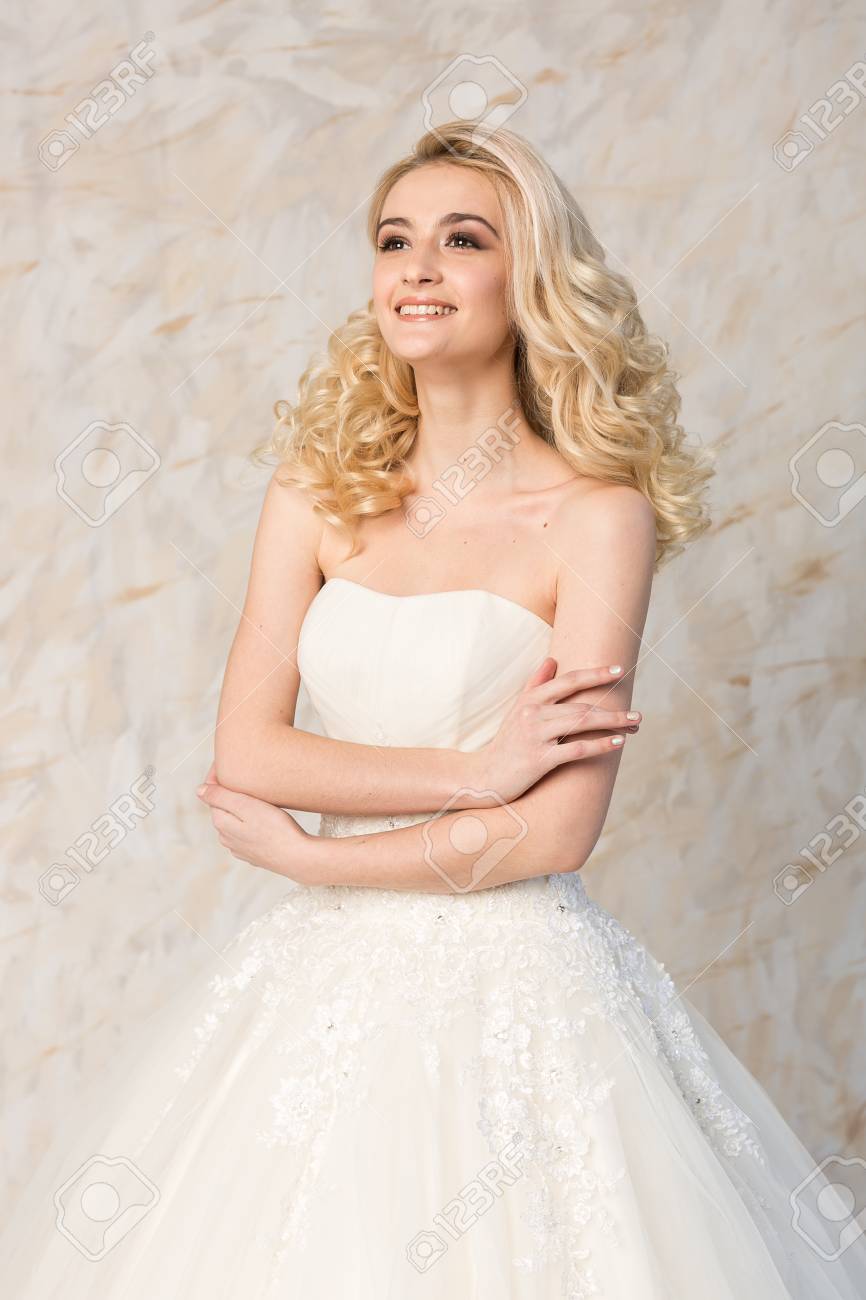 Fashionable White Gown, Beautiful Blonde Model, Bride Hairstyle And Makeup Concept - Young Dreaming Girl In Wedding Festive Dress, Standing Indoors On Light Background, Romantic Slender Woman Posing Stock Photo, Picture And Fashionable White Gown, Beautiful Blonde Model, Bride Hairstyle And Makeup Concept - Young Dreaming Girl In Wedding Festive Dress, Standing Indoors On Light Background, Romantic Slender Woman Posing Stock Photo, Picture And