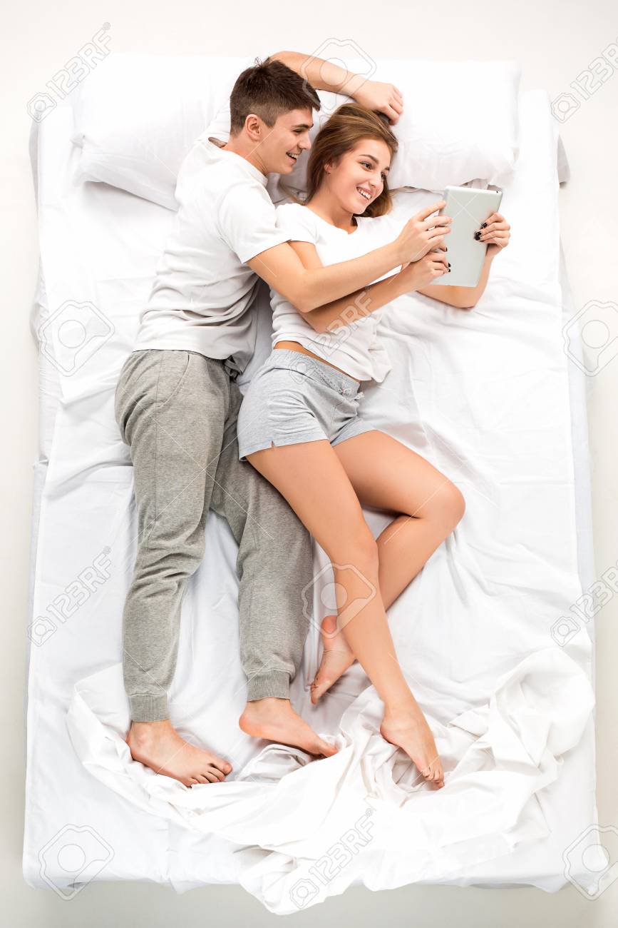 The Young Lovely Couple Lying In A White Bed With Laptop Love