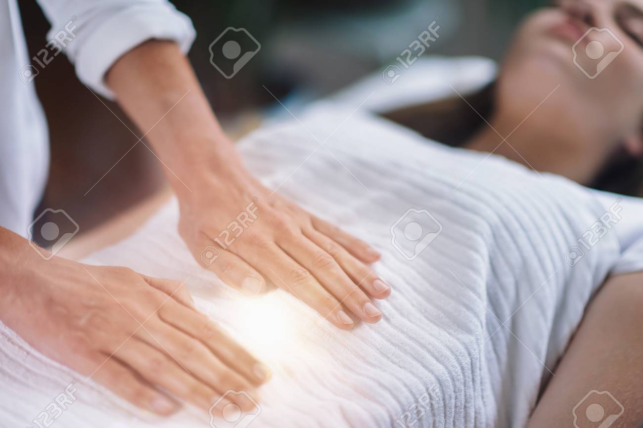 Female therapist performing Reiki therapy treatment holding hands over woman's stomach. Alternative therapy concept of stress reduction and relaxation. - 110034235 Female therapist performing Reiki therapy treatment holding hands over woman's stomach. Alternative therapy concept of stress reduction and relaxation. - 110034235