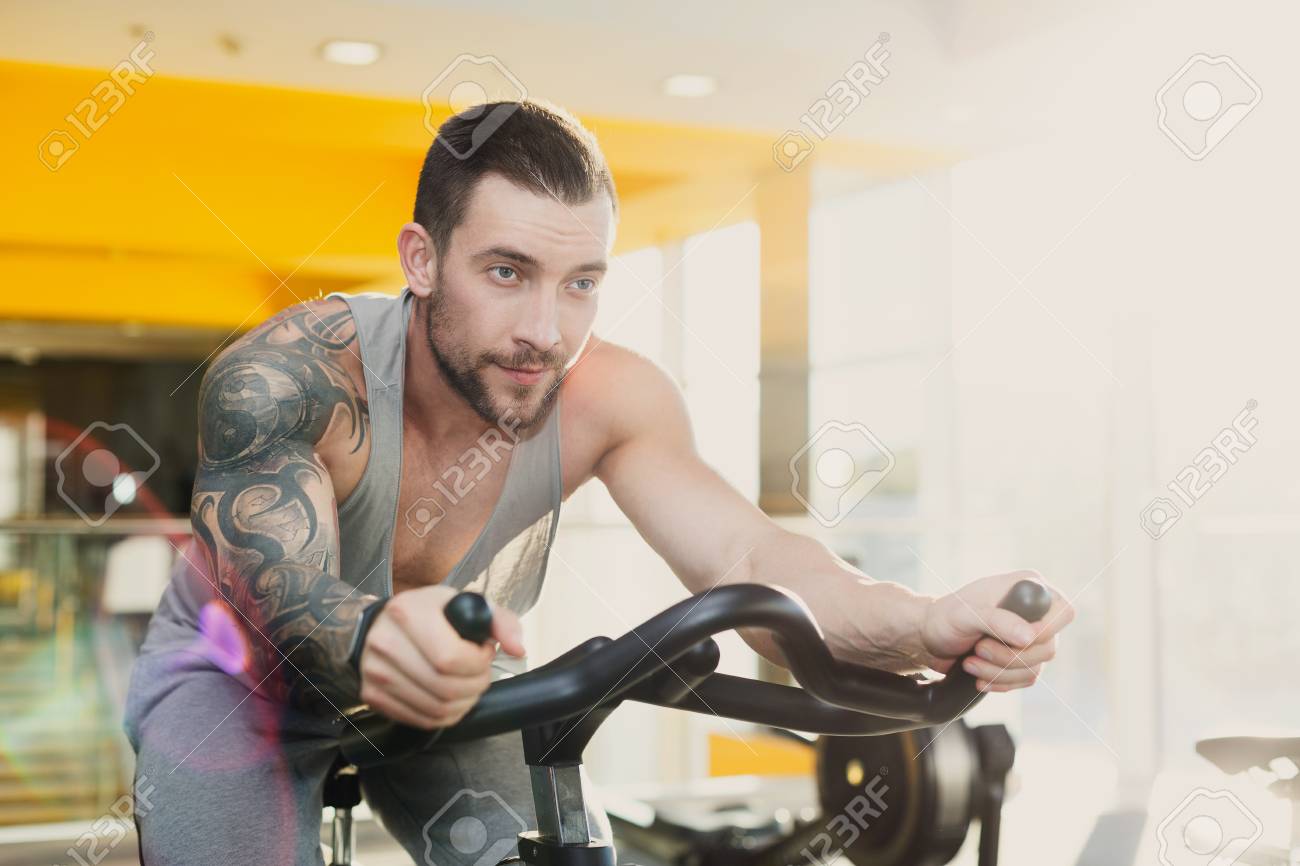 フィットネス クラブのタトゥーを持つ若者 カーディオワーク アウト 乗る文房具自転車 健康的なライフ スタイル 男のジムでトレーニングします フレア効果 の写真素材 画像素材 Image