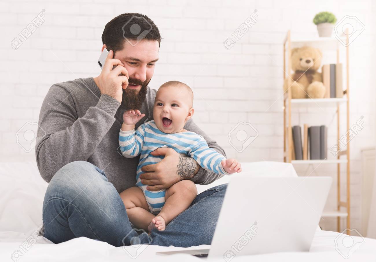 Business at home. Young father talking on mobile phone and nursing newborn child, copy space - 113235446 Business at home. Young father talking on mobile phone and nursing newborn child, copy space - 113235446