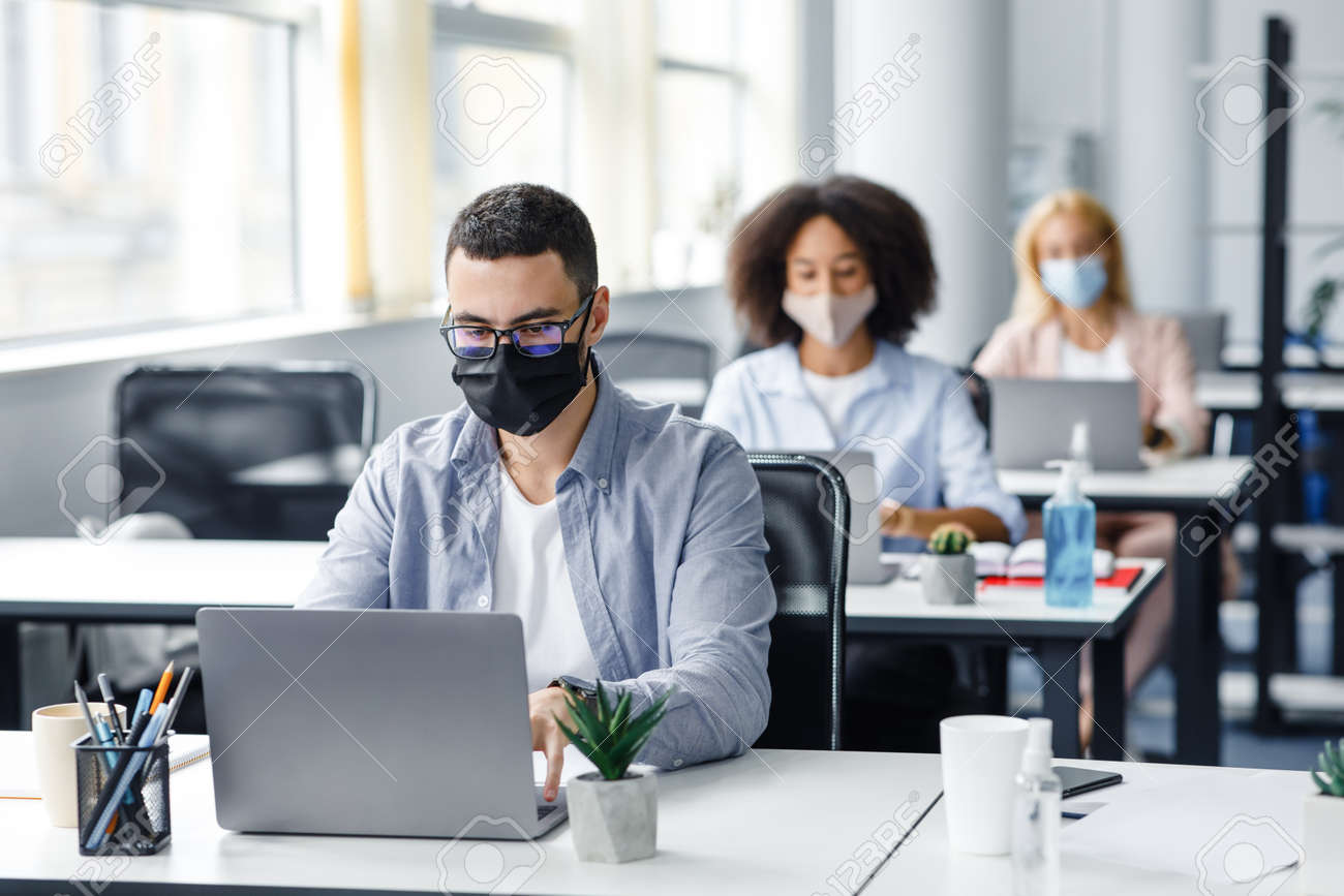 Teamwork in corporate company and returning to work after quarantine covid-19. Focused millennial man in glasses and protective mask works at laptop at workplace with antiseptic in office interior - 156529427 Teamwork in corporate company and returning to work after quarantine covid-19. Focused millennial man in glasses and protective mask works at laptop at workplace with antiseptic in office interior - 156529427