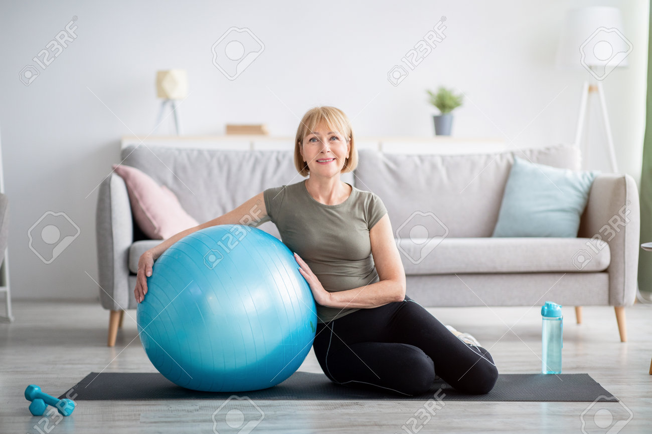 Full length portrait of happy mature woman resting on yoga mat with fitness ball, smiling at camera indoors, free space. Cheerful senior lady taking break from her domestic training - 166099572 Full length portrait of happy mature woman resting on yoga mat with fitness ball, smiling at camera indoors, free space. Cheerful senior lady taking break from her domestic training - 166099572