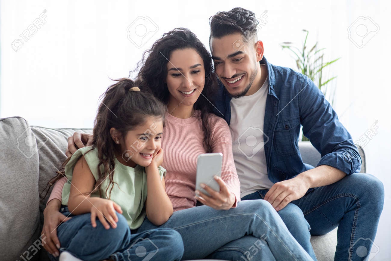 Happy arabic family of three using smartphone at home, relaxing on couch - 165745138 Happy arabic family of three using smartphone at home, relaxing on couch - 165745138