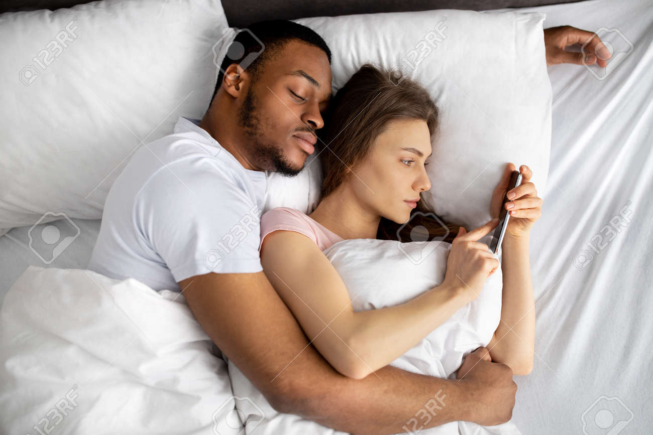 Mujer Joven Acostada En La Cama En Brazos De Su Marido Negro Y Usando El  Teléfono Móvil Para Enviar Mensajes De Texto A Su Amante, Vista Superior  Fotos, retratos, imágenes y fotografía
