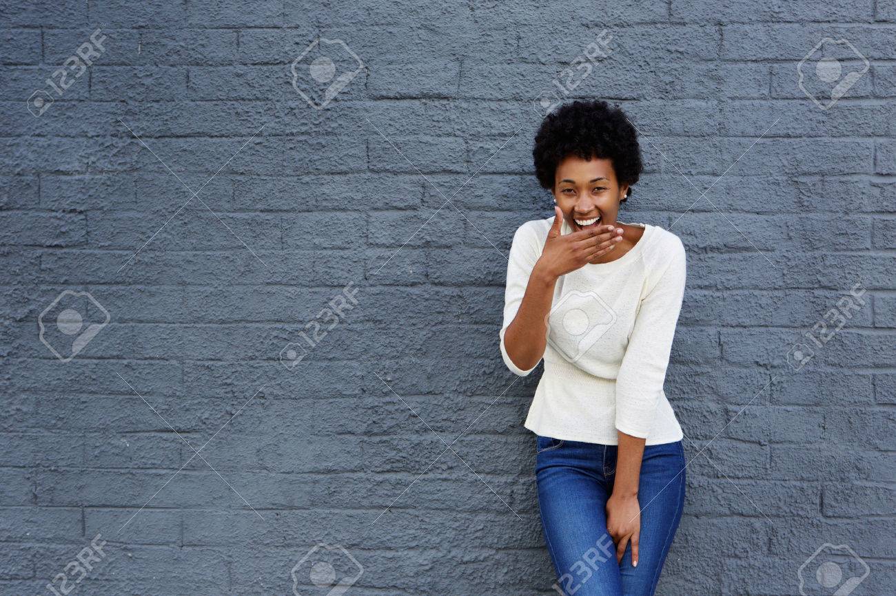 Portrait of happy african woman covering her mouth and laughing against a gray wall - 51497359 Portrait of happy african woman covering her mouth and laughing against a gray wall - 51497359