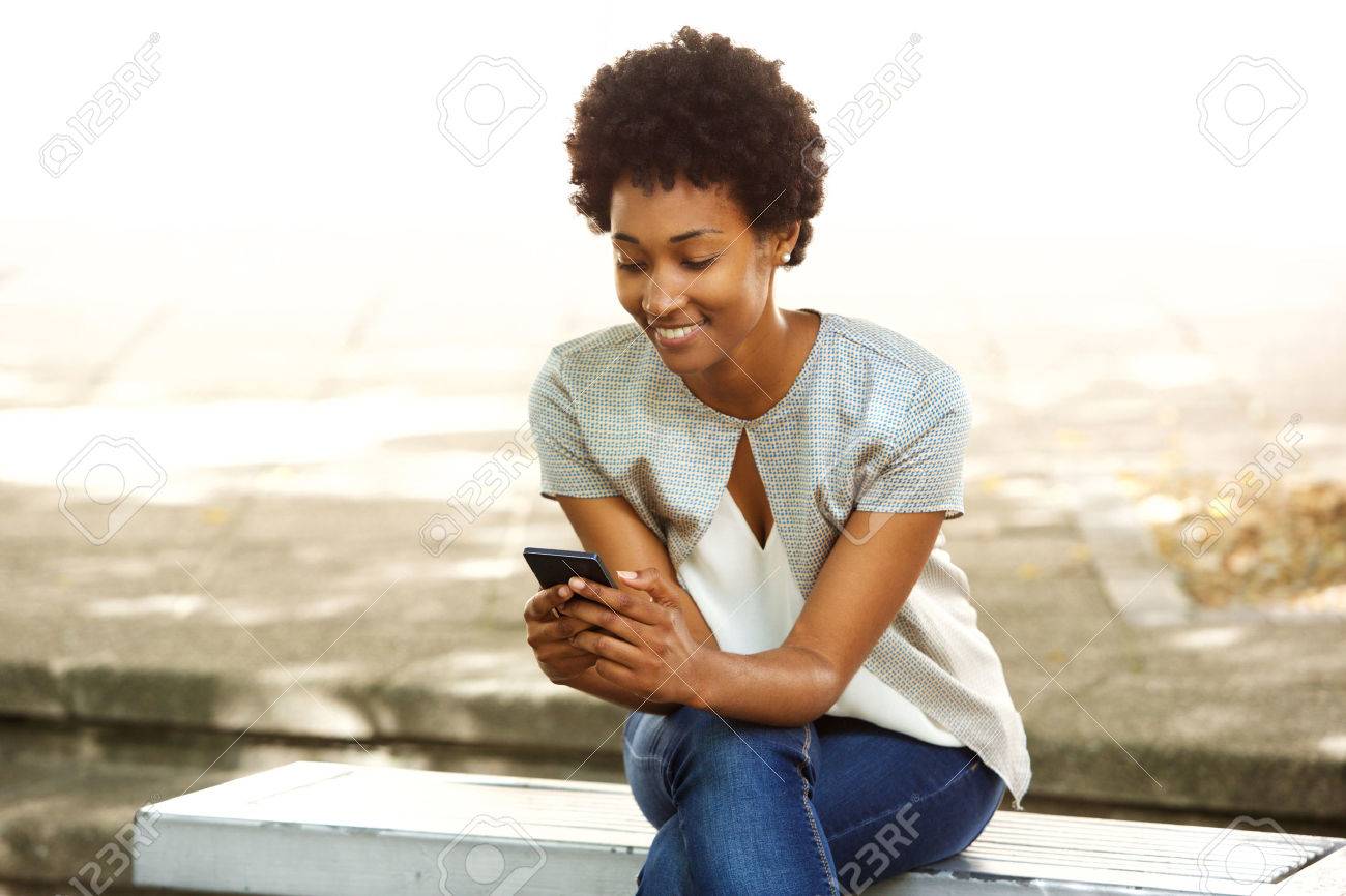 Portrait of happy young african woman sitting outside on bench reading a text message on her mobile phone - 51497505 Portrait of happy young african woman sitting outside on bench reading a text message on her mobile phone - 51497505