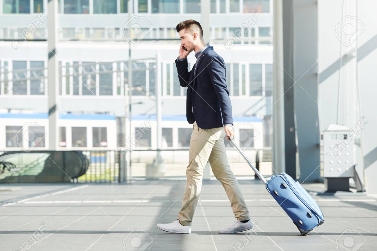 Full length side portrait of business man walking with luggage on telephone call at station - 68850459 Full length side portrait of business man walking with luggage on telephone call at station - 68850459