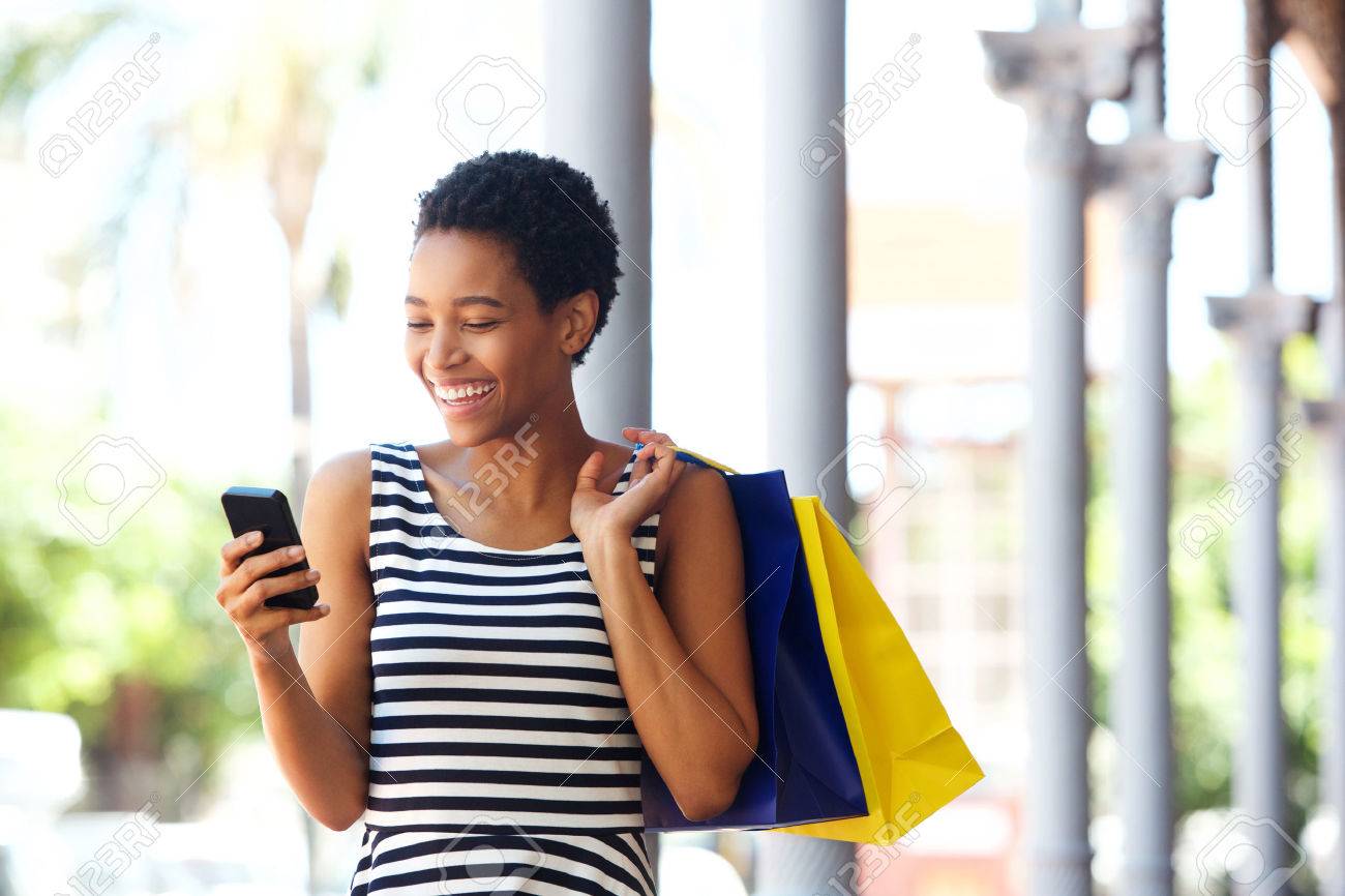 Portrait of happy young african american woman walking with cellphone and shopping bags - 75916537 Portrait of happy young african american woman walking with cellphone and shopping bags - 75916537