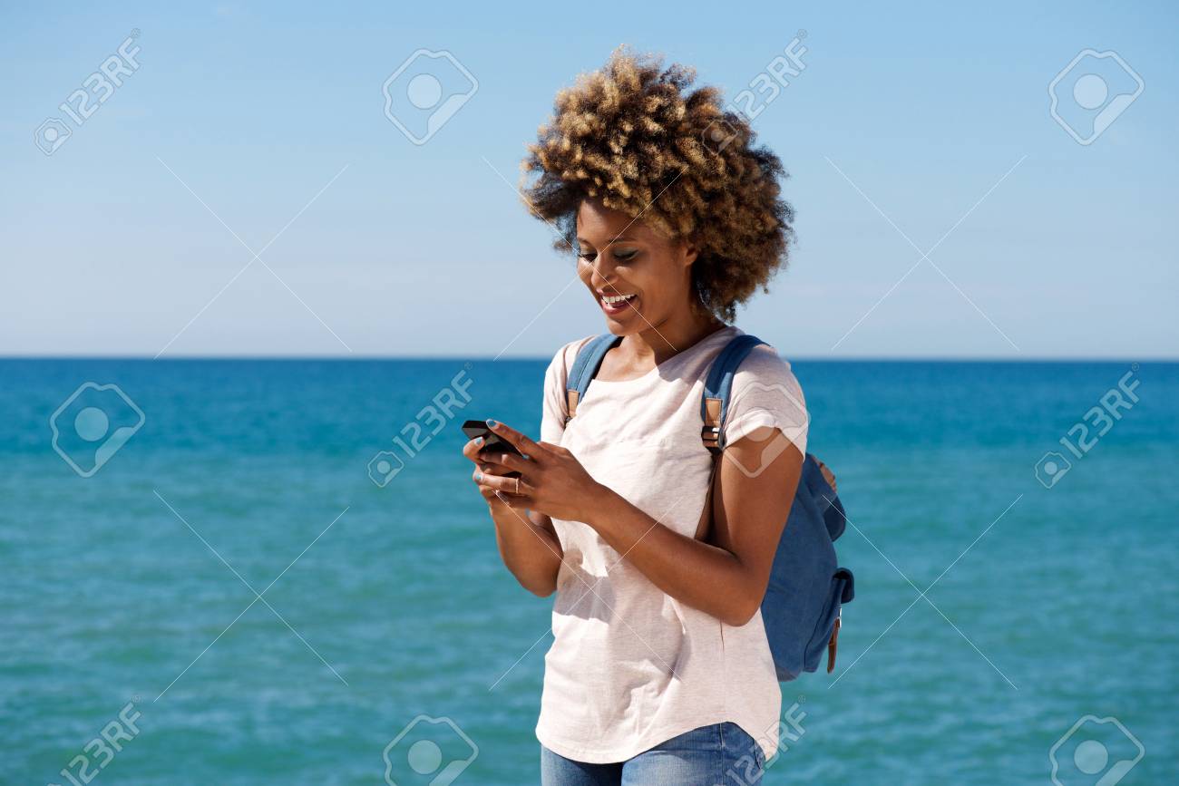 Portrait of smiling african woman on the beach reading text message on smart phone - 85260980 Portrait of smiling african woman on the beach reading text message on smart phone - 85260980