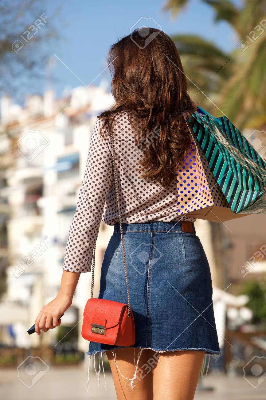 Rear portrait of woman walking in jean skirt with gift bags and cellphone - 93545470 Rear portrait of woman walking in jean skirt with gift bags and cellphone - 93545470