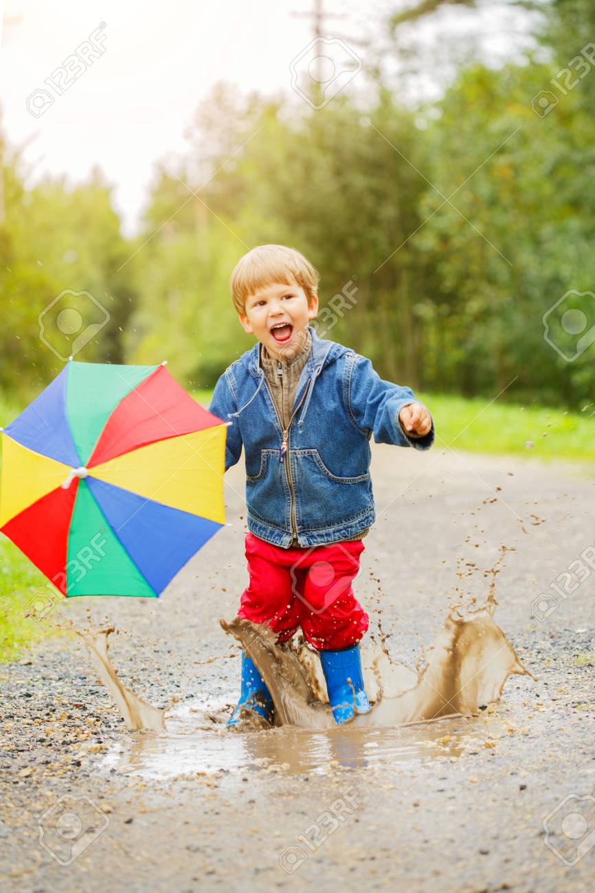 El Salta En Charcos Con Botas. En La Lluvia Un Niño Con Un De Arco Iris Está Caminando Afuera. Otoño, Vacaciones Fotos, Retratos, Y Fotografía De Archivo Libres
