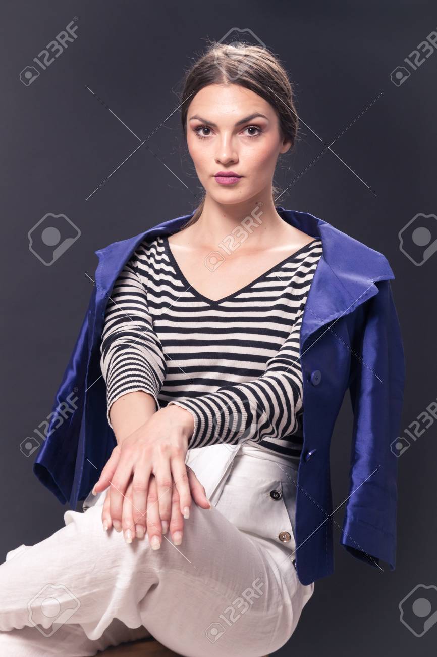 一人の若い白人女性 代 29 歳 スツール ポーズ スタジオ 白い背景 ファッショナブルな青いジャケット 水平方向のストライプ シャツ バーに座っているファッション モデル ホワイト パンツ カメラを の写真素材 画像素材 Image