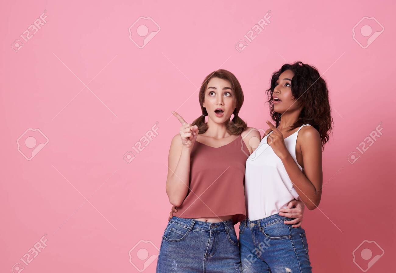 Portrait of two shocked young woman standing with her finger pointing at copy space isolated over pink banner background. - 130127768 Portrait of two shocked young woman standing with her finger pointing at copy space isolated over pink banner background. - 130127768