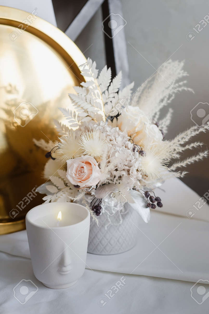 Stylish composition of preserved flowers and dried flowers in a light color scheme on the background of a golden tray with a burning candle. Photo of a bouquet with roses in the interior. - 170743101 Stylish composition of preserved flowers and dried flowers in a light color scheme on the background of a golden tray with a burning candle. Photo of a bouquet with roses in the interior. - 170743101