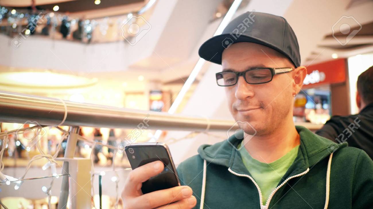 キャップと黒縁メガネを身に着けている若い白人男が彼の携帯電話のタッチ スクリーンをタップします の写真素材 画像素材 Image