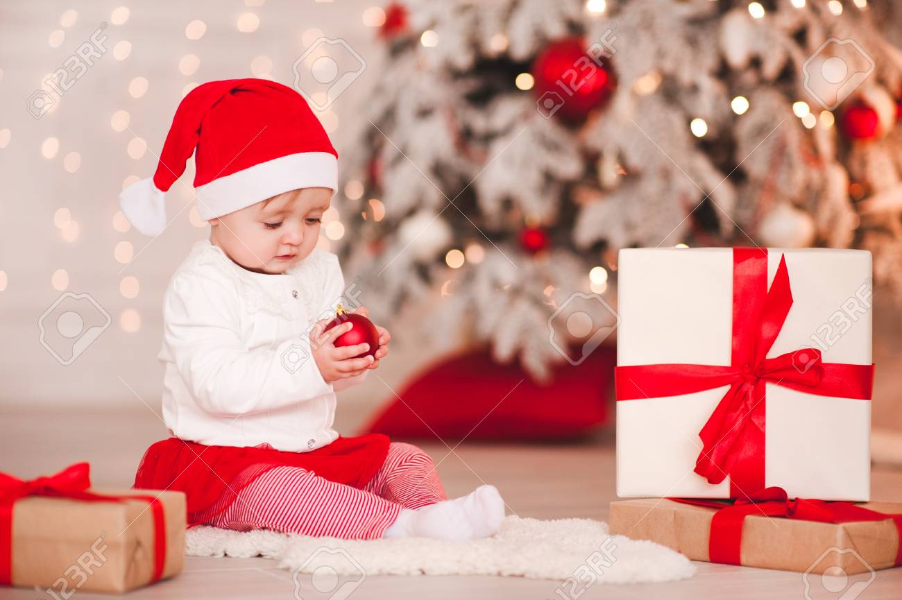 かわいい女の子1歳は クリスマスの背景の上にポーズサンタ帽子を身に着けています クリスマスボールで床に座って ホリデーシーズン の写真素材 画像素材 Image