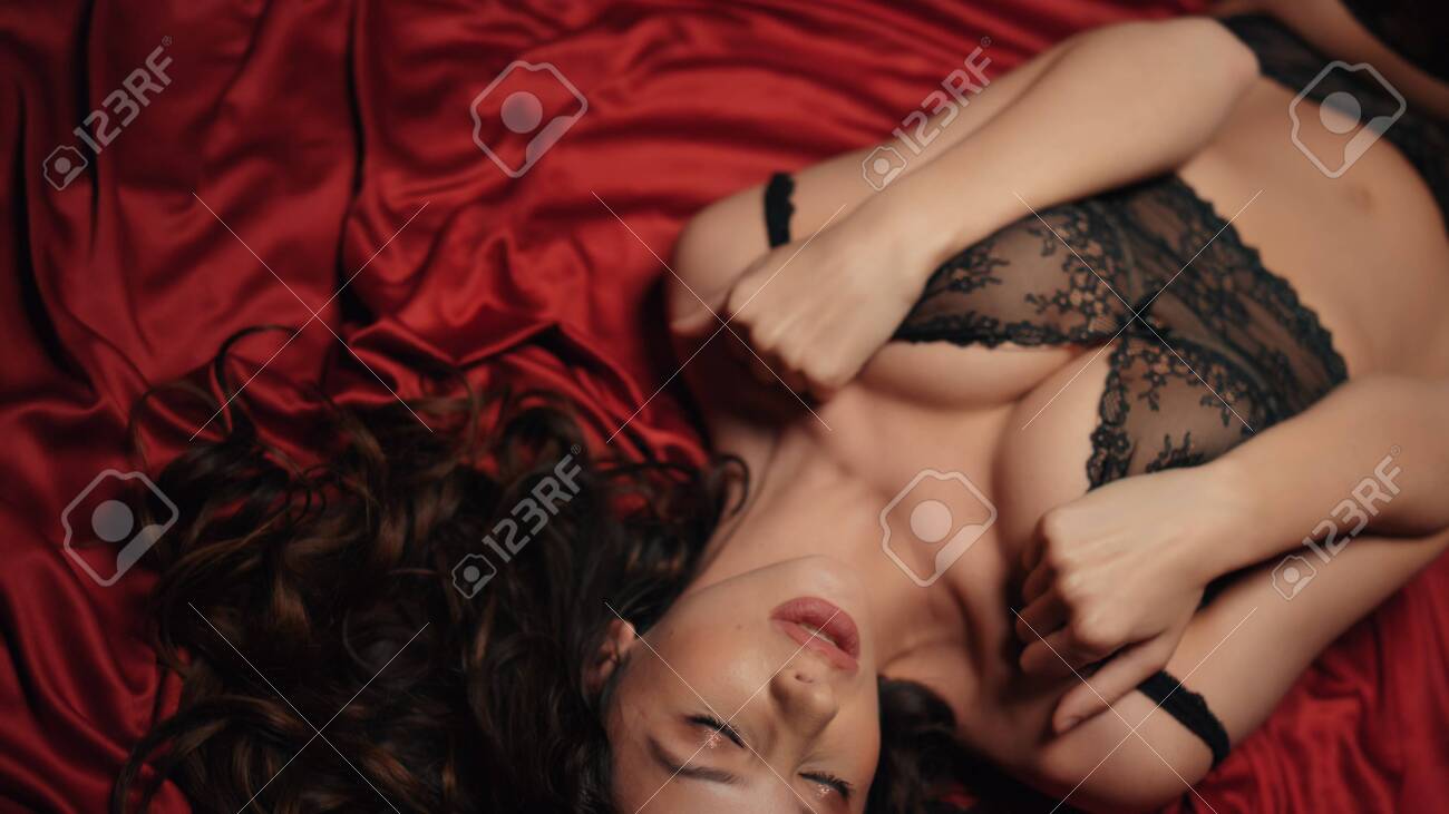 Top View Sexy Woman Squeezing Breast In Lace Bra On Red Silk Cloth.  Passionate Girl Touching Body In Black Lingerie And Stockings On Bed.  Seductive Model Girl Lying On Satin Sheets Indoors.