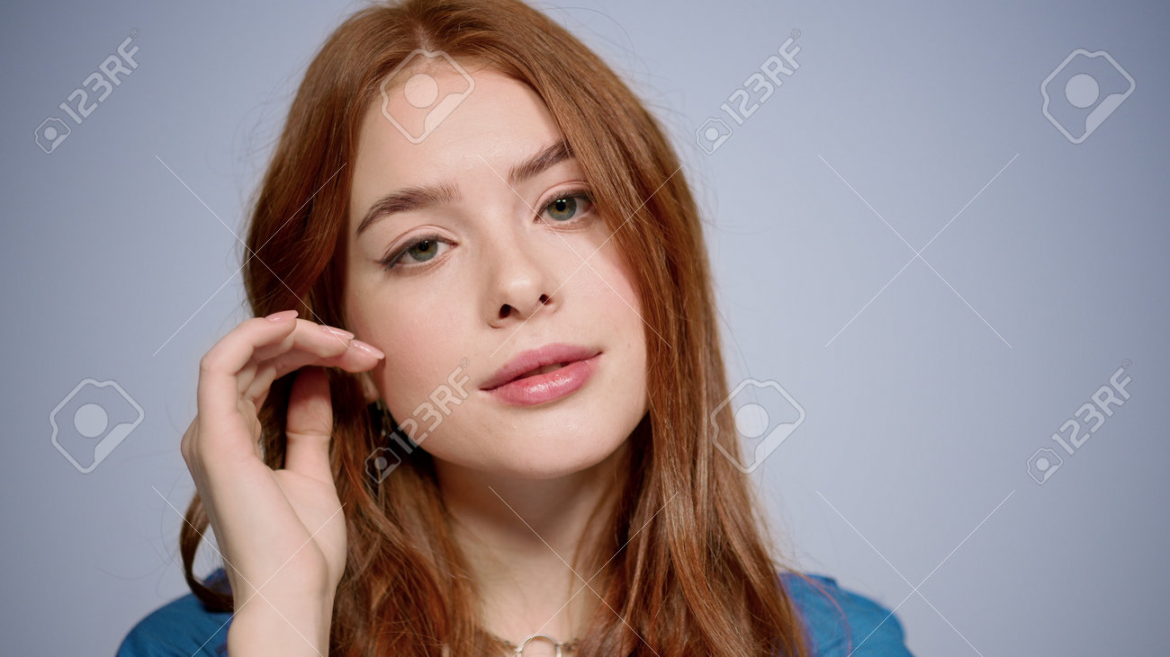 Pretty red hair girl flirting in studio in slow motion. Closeup romantic woman touching face indoors. Portrait of gorgeous female person looking at camera on gray background. - 161621022 Pretty red hair girl flirting in studio in slow motion. Closeup romantic woman touching face indoors. Portrait of gorgeous female person looking at camera on gray background. - 161621022