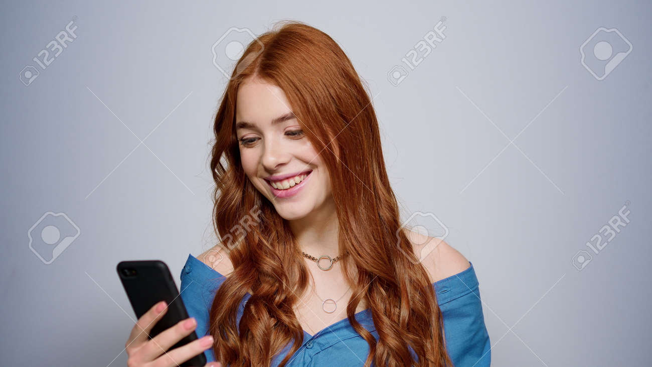 Red hair girl holding mobile phone in studio. Portrait of cheerful female person looking smartphone screen indoors. Closeup attractive woman getting good news on cellphone. - 159609837 Red hair girl holding mobile phone in studio. Portrait of cheerful female person looking smartphone screen indoors. Closeup attractive woman getting good news on cellphone. - 159609837