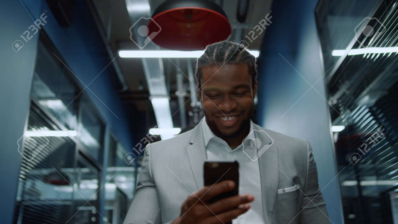 Portrait of smiling afro business man getting good news on mobile phone in office. Closeup happy african businessman reading message on smartphone in corridor. African american man laughing in hallway - 158485725 Portrait of smiling afro business man getting good news on mobile phone in office. Closeup happy african businessman reading message on smartphone in corridor. African american man laughing in hallway - 158485725