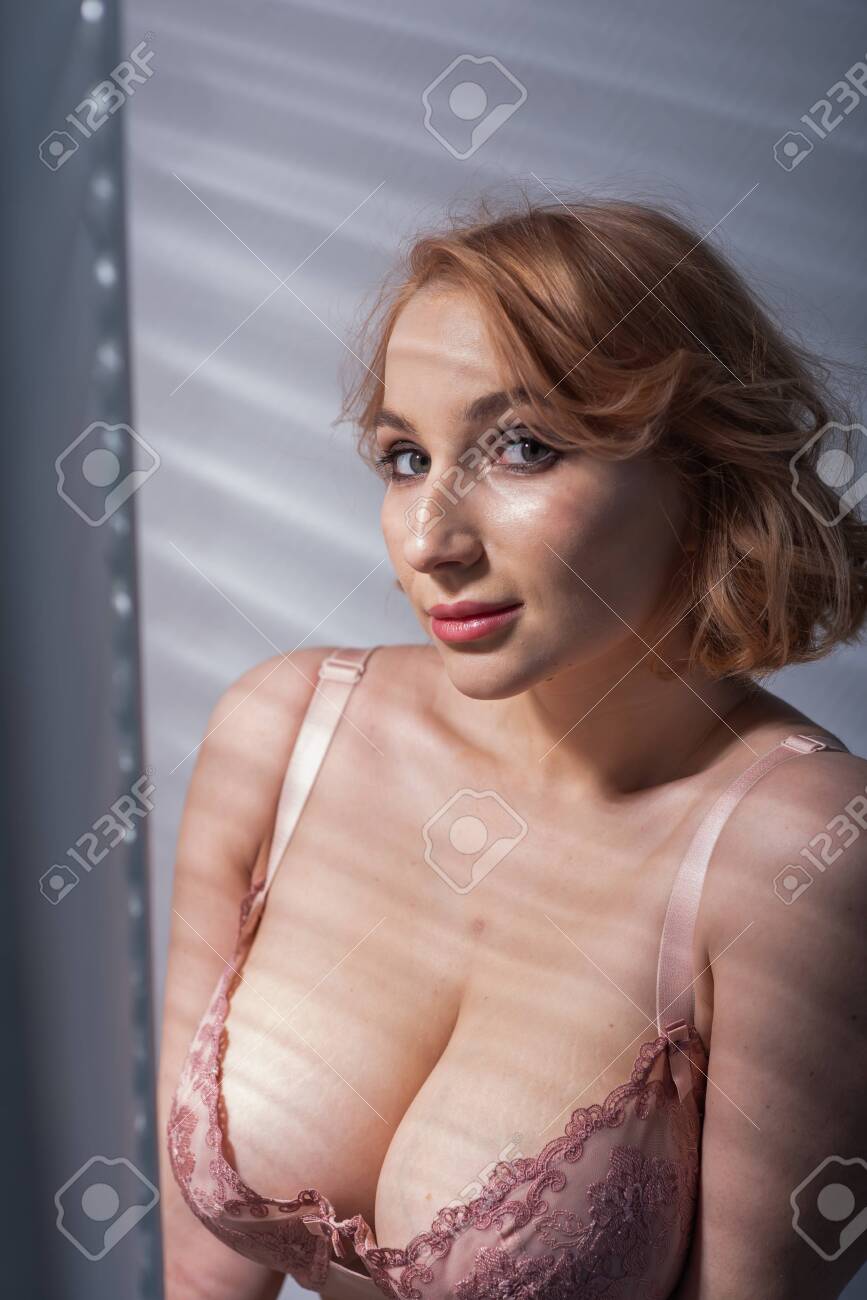 A Beautiful Young Woman With Very Large Natural Breasts Is Standing By The  Window With Shutters. A Girl In Pink Lace Shows A Beautiful Figure Next To  A White Screen. Huge Female