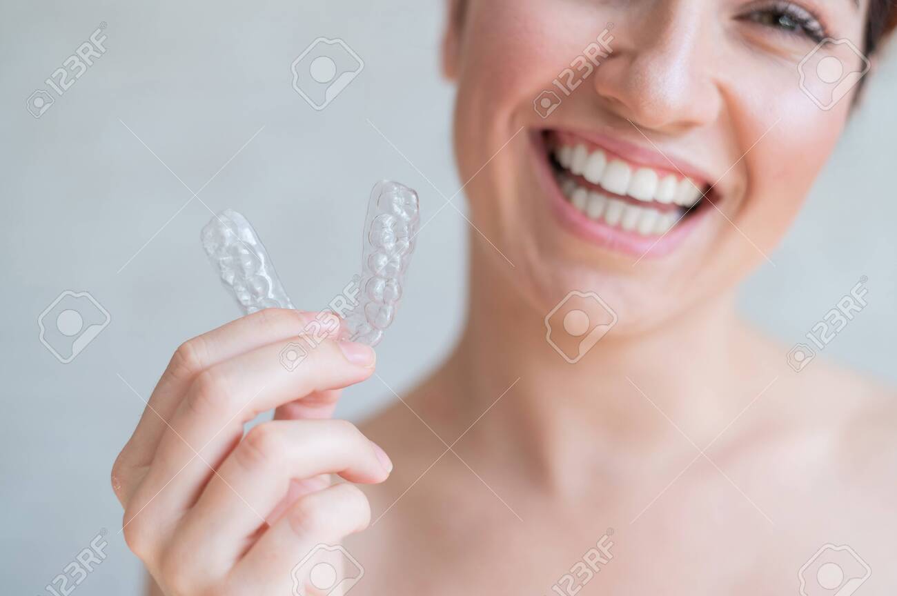 Close-up of orthodontic silicone transparent teeth aligner in female hands. A woman with a perfect charm smile holds a removable night retainer. Bracket for teeth whitening. Cropped photo. - 148270945 Close-up of orthodontic silicone transparent teeth aligner in female hands. A woman with a perfect charm smile holds a removable night retainer. Bracket for teeth whitening. Cropped photo. - 148270945