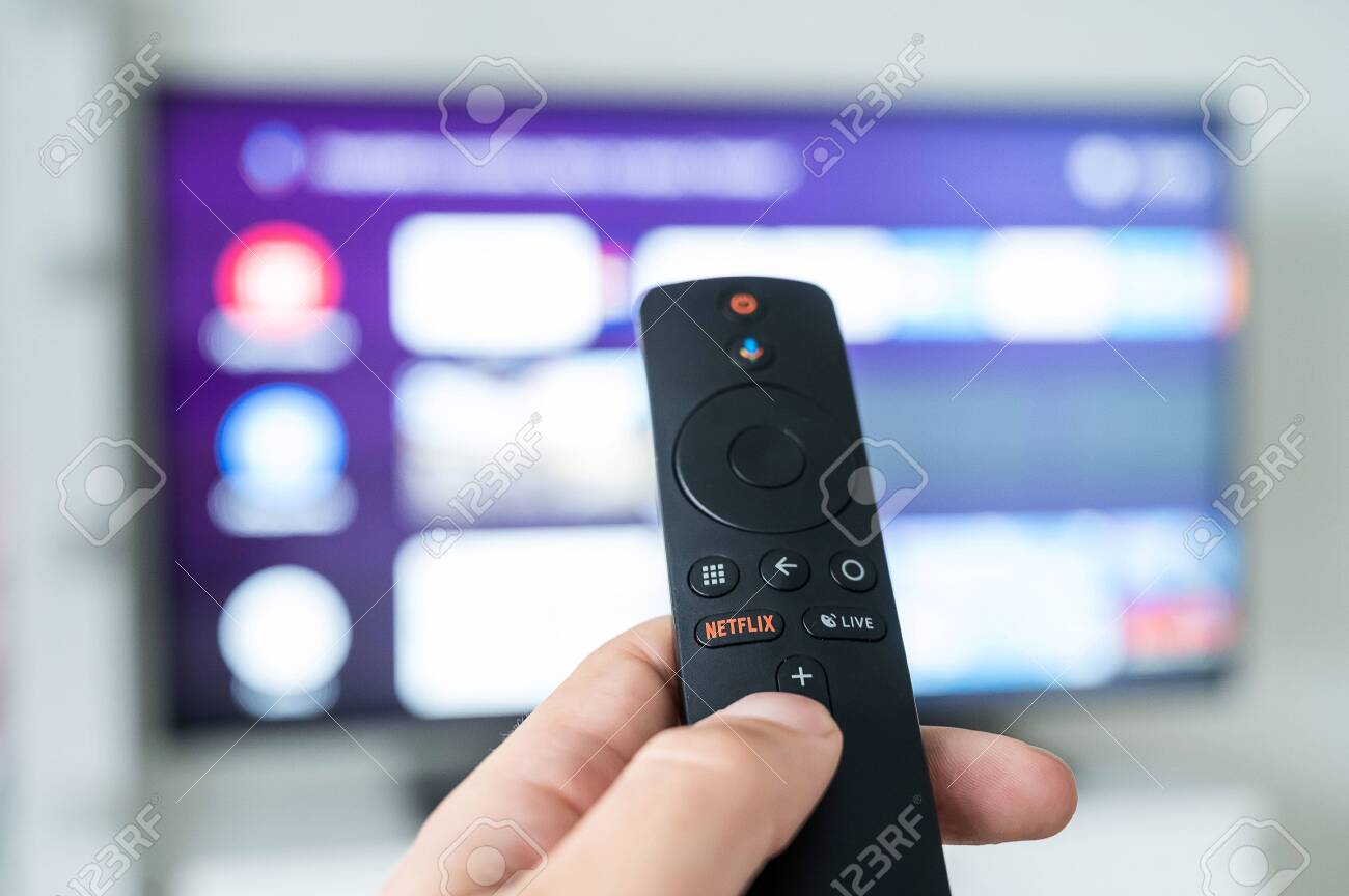 한 남자가 흐릿한 텔레비전 배경에 넷플릭스 로고가 있는 리모콘을 손에 들고 있습니다. 영화 및 TV 프로그램에 무제한으로 액세스할 수  있는 포털. 로열티 무료 사진, 그림, 이미지 그리고 스톡포토그래피. Image 151934126