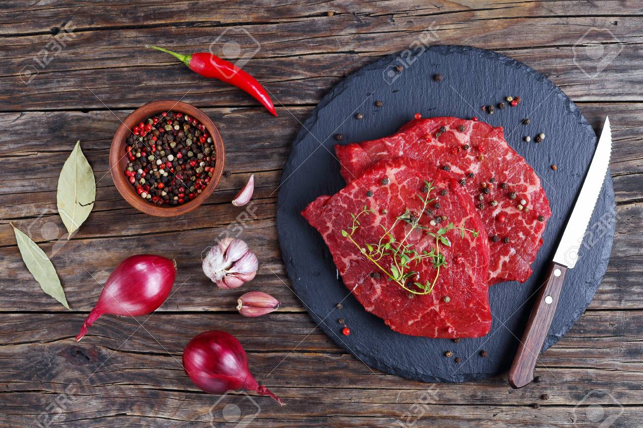 2 つの生の霜降り牛ステーキ油に注がれ 上からの眺めナイフ スパイス タマネギ 古い木製のテーブルに月桂樹の葉と黒いスレート トレイに緑 新鮮なタイムの 葉をトッピング クローズ アップ の写真素材 画像素材 Image