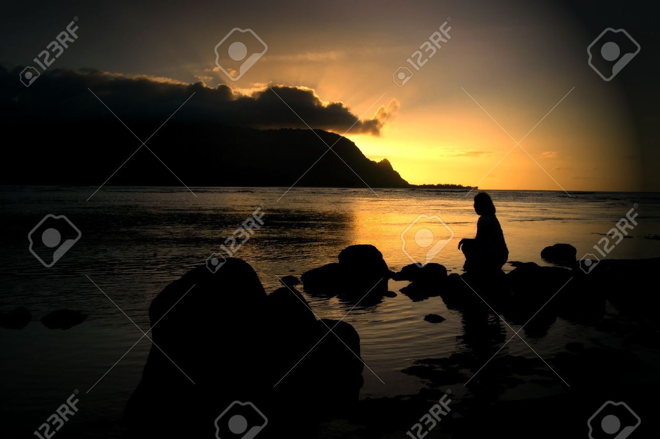 Woman sits alone on a beach at Princeville, Kauai and watches the sun set behind a bank of clouds and the Kauai mountains. Sunset tints the sky and waters a brilliant gold. - 14922799 Woman sits alone on a beach at Princeville, Kauai and watches the sun set behind a bank of clouds and the Kauai mountains. Sunset tints the sky and waters a brilliant gold. - 14922799