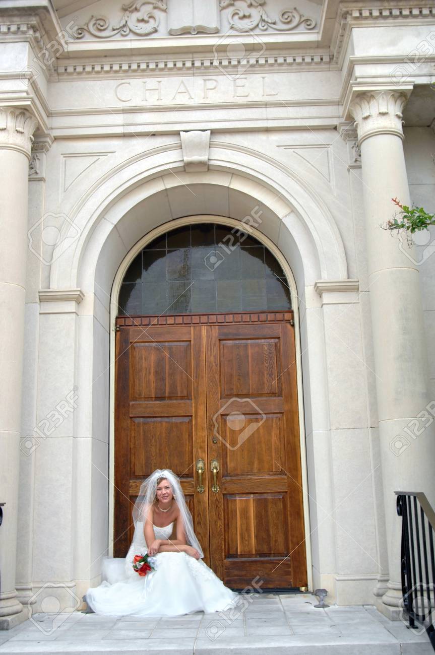 花嫁は結婚式のチャペルのドアでひざまずきます 彼女は笑みを浮かべて 彼女の赤とオレンジのバラの花を保持します エレガントな建物は彼女の頭の上に刻まれた チャペル の写真素材 画像素材 Image