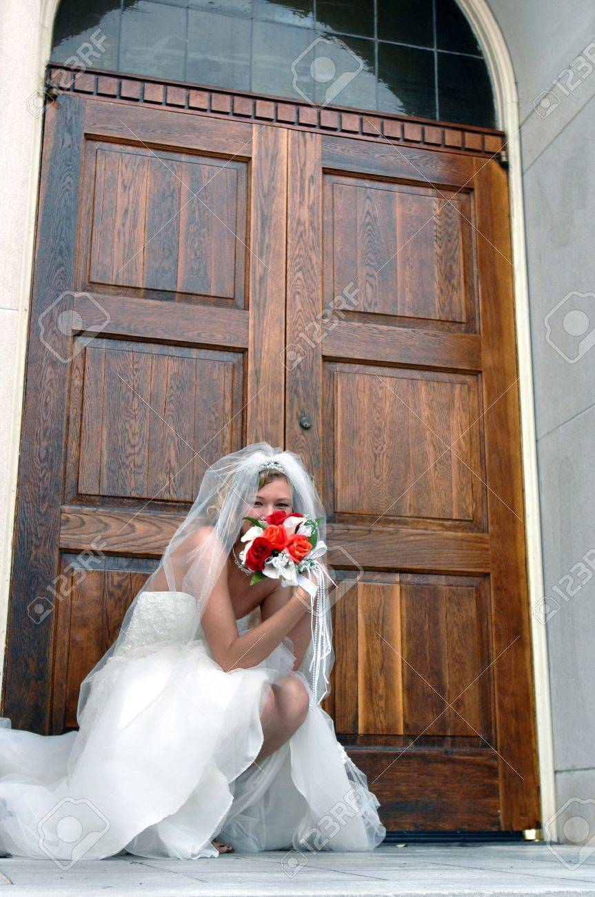 Bride kneels besides the chapel door, anticipating the doors opening for the ceremony She is teasingly hiding her face behind he bouquet of red and orange roses - 15024480 Bride kneels besides the chapel door, anticipating the doors opening for the ceremony She is teasingly hiding her face behind he bouquet of red and orange roses - 15024480