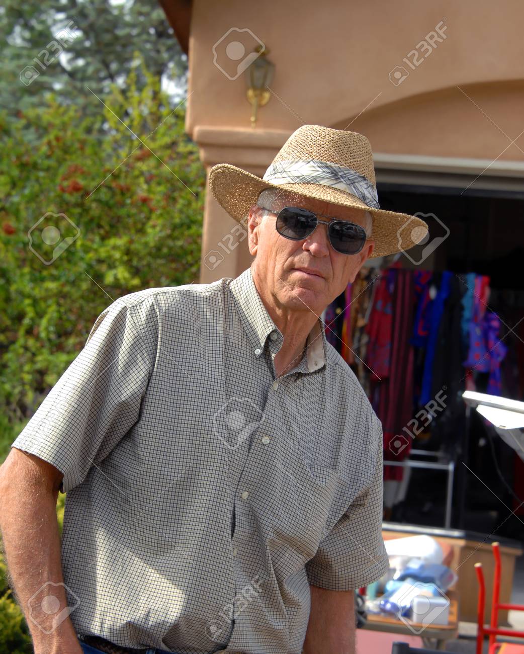Hombre Maduro Prepara En Sus Compras Para Mirar La Cámara. Está Tomando El Café De Descanso Y Unas De Sol De La Paja Y La Camisa De Estilo De
