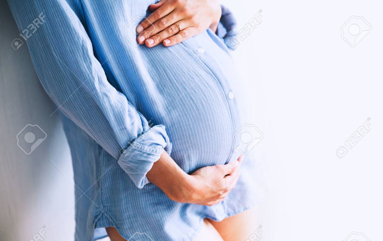 Pregnant woman holds hands on belly at home interiors. Pregnancy, parenthood, preparation and expectation concept. Close-up, copy space, indoors. - 76412878 Pregnant woman holds hands on belly at home interiors. Pregnancy, parenthood, preparation and expectation concept. Close-up, copy space, indoors. - 76412878
