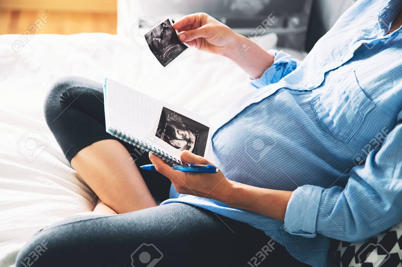 Pregnant woman makes notes in notebook and holding ultrasound image and medical documents at home interiors. Pregnancy, parenthood, preparation and expectation concept. Close-up, copy space, indoors. - 77855984 Pregnant woman makes notes in notebook and holding ultrasound image and medical documents at home interiors. Pregnancy, parenthood, preparation and expectation concept. Close-up, copy space, indoors. - 77855984