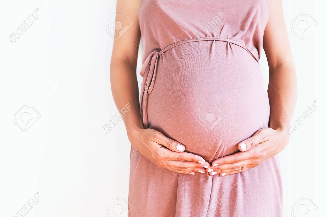 Pregnant woman in dress holds hands on belly on a white background. Pregnancy, maternity, preparation and expectation concept. Close-up, copy space, indoors. Beautiful tender mood photo of pregnancy. - 84179244 Pregnant woman in dress holds hands on belly on a white background. Pregnancy, maternity, preparation and expectation concept. Close-up, copy space, indoors. Beautiful tender mood photo of pregnancy. - 84179244
