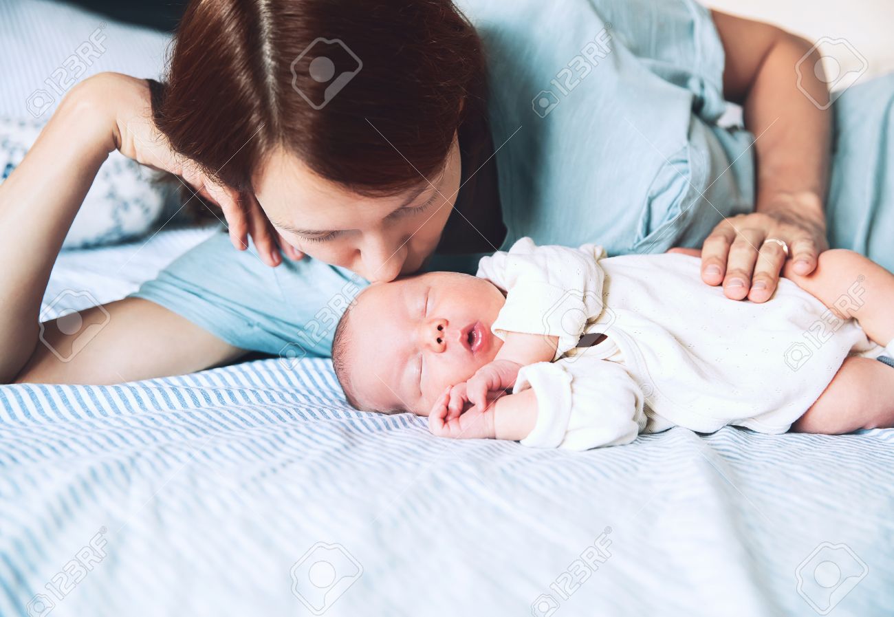 Mother kissing her newborn baby. Young beautiful mom lying at bed with a cute little sleeping child. New born baby's first days of life in family at home. Loving mother looks at asleep infant. - 86515086 Mother kissing her newborn baby. Young beautiful mom lying at bed with a cute little sleeping child. New born baby's first days of life in family at home. Loving mother looks at asleep infant. - 86515086